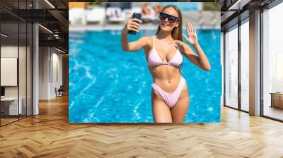 Portrait of a happy beautiful woman in bikini and sunglasses making selfie photo on smartphone wave hello for social media at pool Wall mural